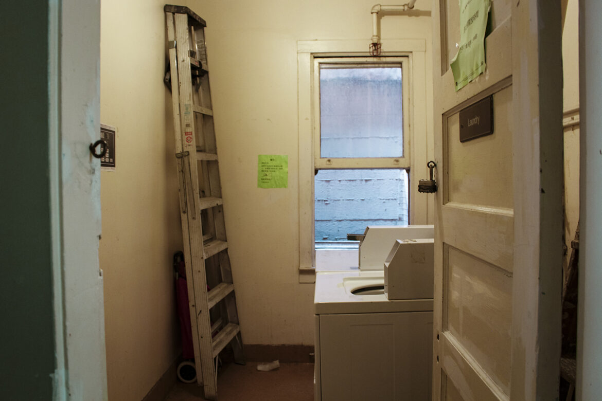 A ladder is propped up in front of a washer and dryer unit in a single-room occupancy building. A long window in the back of the room is partially open.