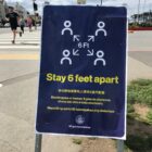 A poster with a blue background and white and yellow graphics and lettering placed near a sidewalk urges people to "Stay 6 feet apart."